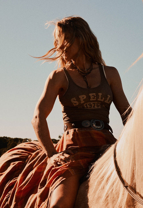 A woman riding a horse outdoors at sunset, wearing a green tank top with 'SPELL 1976' printed on the front, a rust-orange plaid skirt, and a large silver belt buckle.