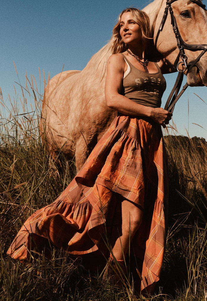 A woman in a rust-orange plaid maxi skirt stands outdoors in tall grass beside a light-colored horse, holding its reins under warm golden sunlight.