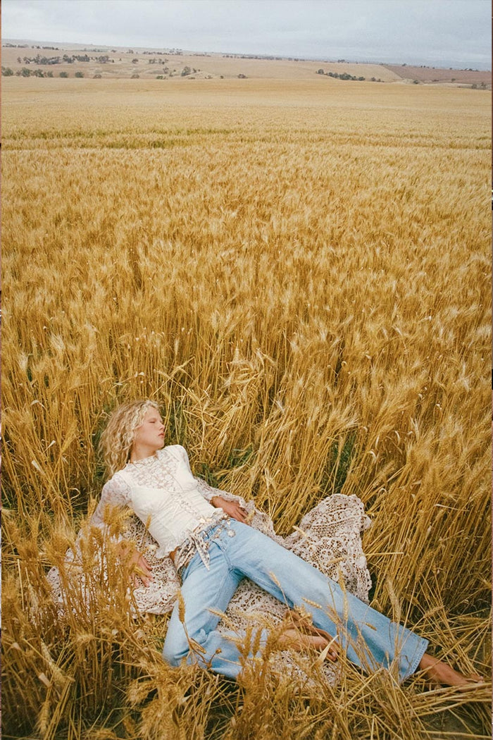 Model lying in a wheat field wearing vintage blue organic cotton relaxed wide-leg jeans styled with a white lace top