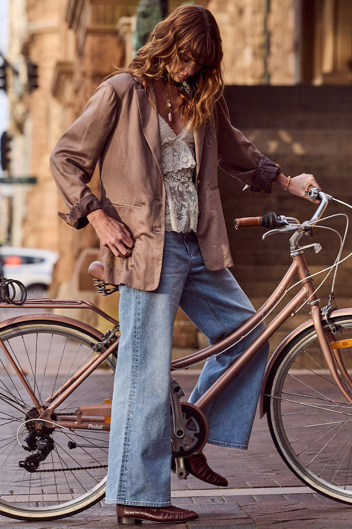 Model standing beside a vintage bicycle, wide-leg Vintage Blue jeans styled with brown satin blazer and lace cami