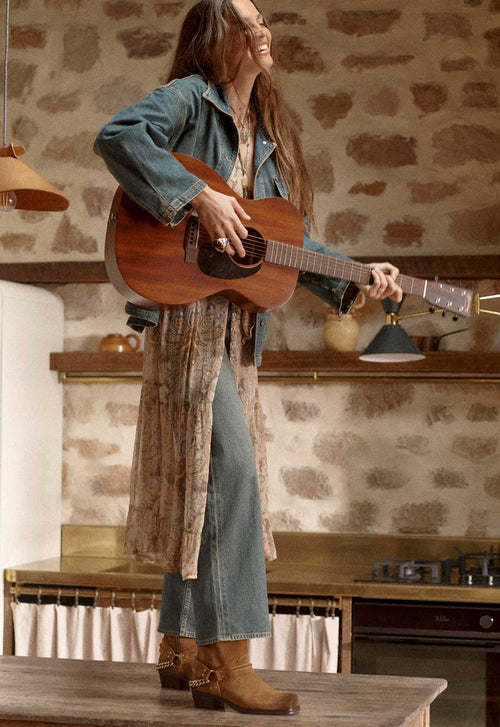 A woman stands in a rustic kitchen setting, holding and playing an acoustic guitar. She is wearing a blue denim jacket, blue jeans, and brown leather boots.