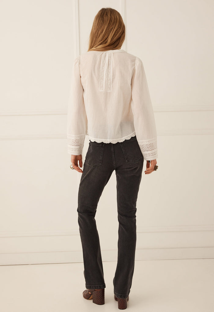 A woman viewed from behind wearing a cream-colored long-sleeve blouse with lace trim and dark charcoal gray jeans, standing against a white paneled wall.