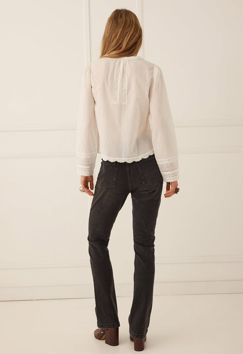 A woman viewed from behind wearing a cream-colored long-sleeve blouse with lace trim and dark charcoal gray jeans, standing against a white paneled wall.