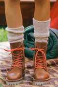 Brown boots with orange laces worn with white socks on a patterned surface.