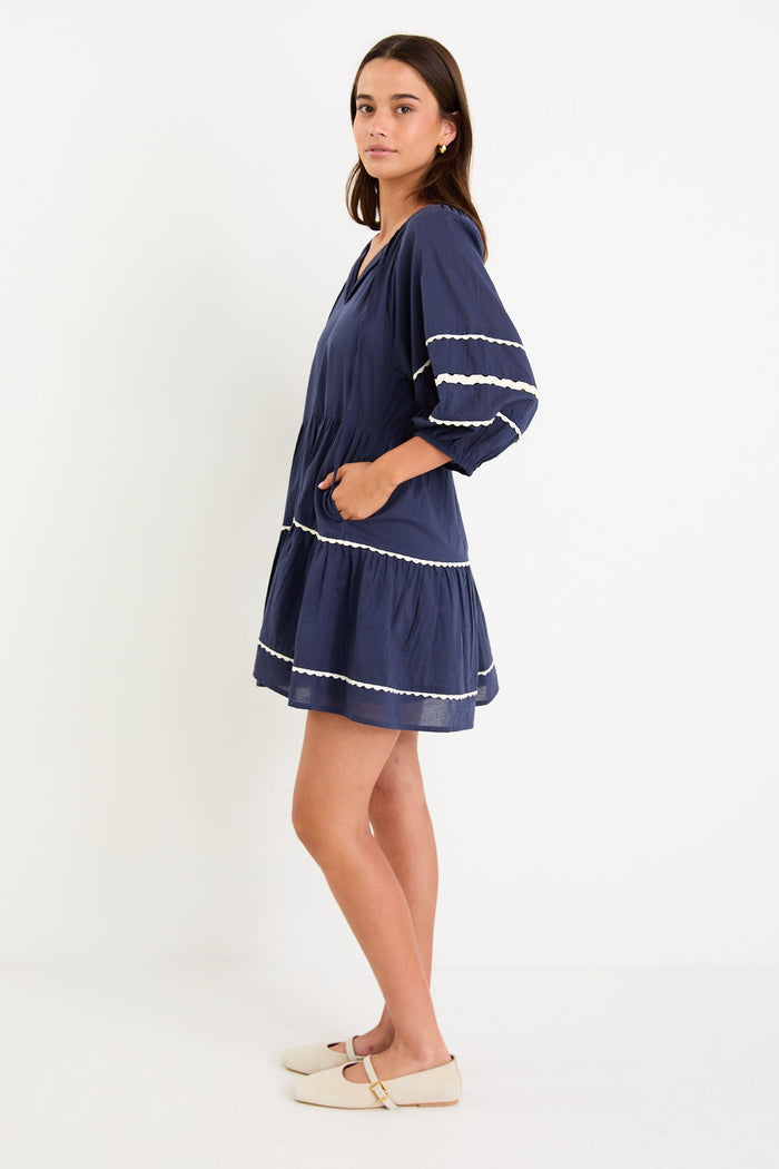 Model wearing a navy tiered short sleeve smock dress with a relaxed fit and contrast trim detail.
