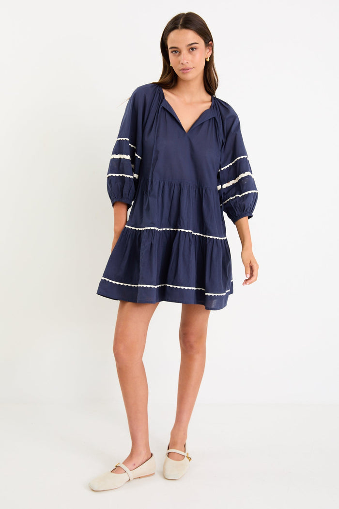 Model wearing a navy tiered short sleeve smock dress with a relaxed fit and contrast trim detail.