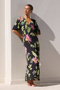 A woman wearing a black maxi dress with a vibrant pink and green tropical floral print stands barefoot by a swimming pool against a white wall.