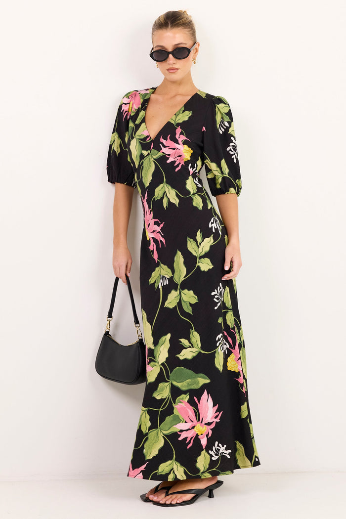 A woman modeling a black maxi dress with a vibrant pink and green floral print, styled with a black handbag and sunglasses against a plain white studio background.