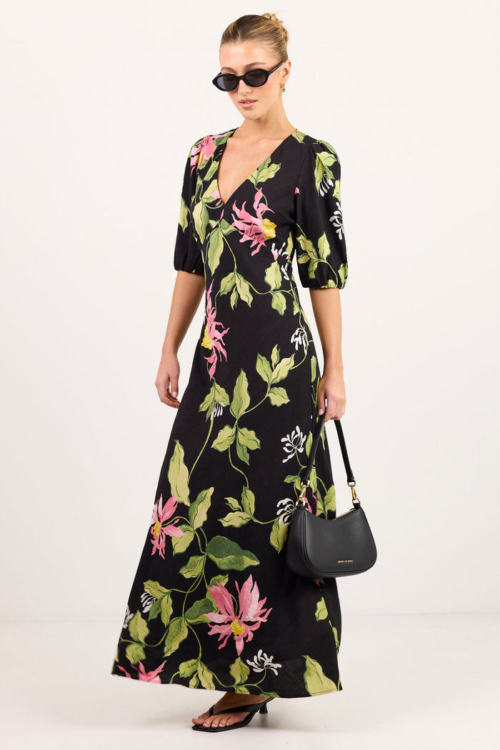 A woman modeling a black maxi dress with a vibrant pink and green floral print, styled with black sunglasses, brown sandals, and a black handbag against a plain white studio background.