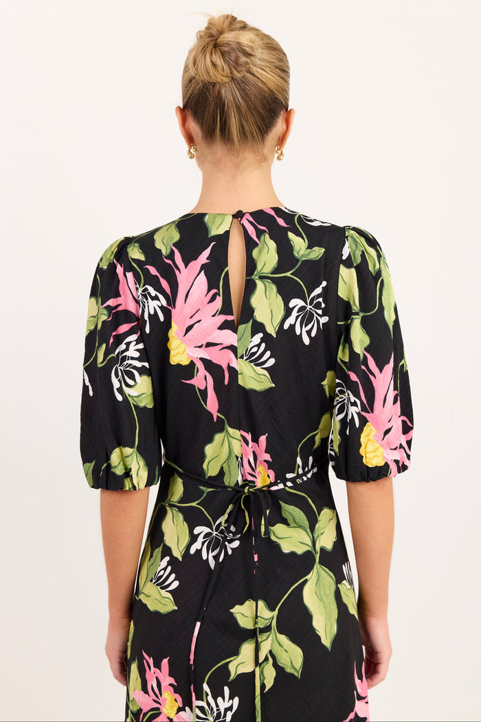 A woman viewed from the back wearing a black dress with a vibrant pink, green, and yellow floral print. The dress features puff sleeves, a tie waist, and a small keyhole cutout at the back of the neck, set against a plain off-white background.
