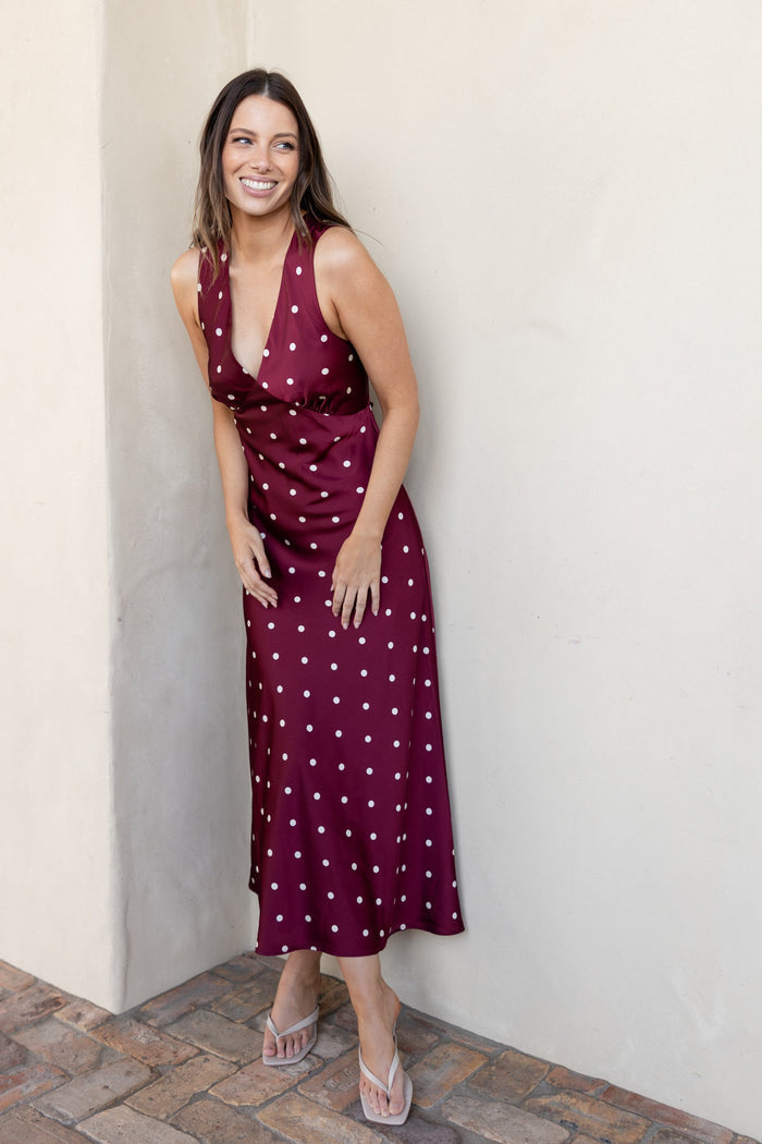 A woman wearing a sleeveless burgundy midi dress with white polka dots, standing against a beige stucco wall on a brick patio.