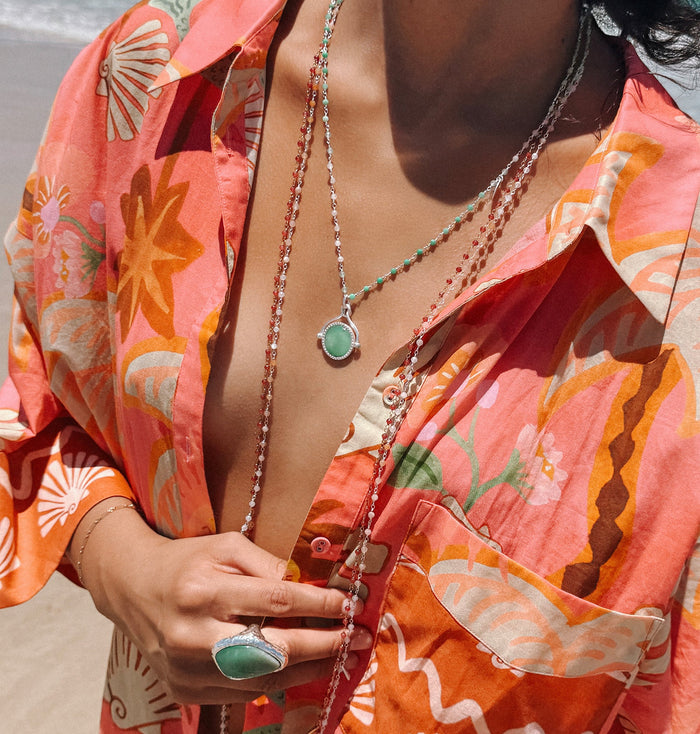 Layered beaded necklaces with green stone pendant and ring styled on floral shirt, shot at beach