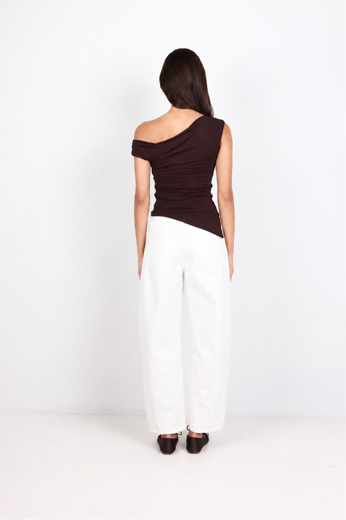 A woman viewed from behind wearing a dark brown off-the-shoulder ruched top and white wide-leg pants, standing against a plain white studio background.