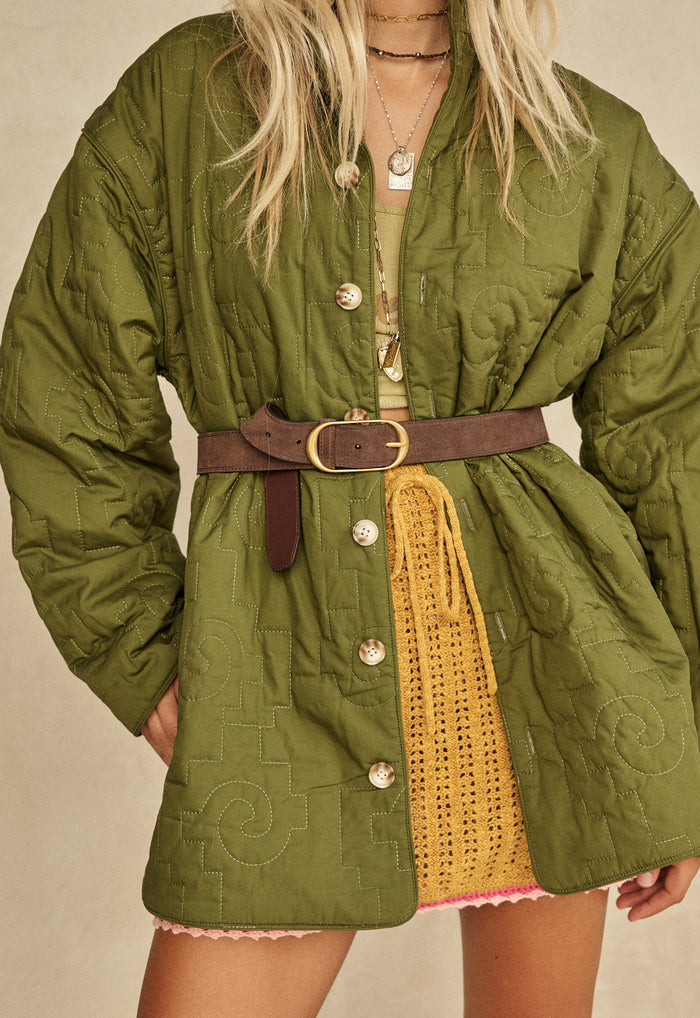 Front detail of mustard knit mini skirt styled under olive quilted jacket belted at waist, embroidery flower design visible