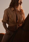 A woman wearing a tan button-up shirt tied at the waist and brown shorts, riding a horse in warm golden-hour light.