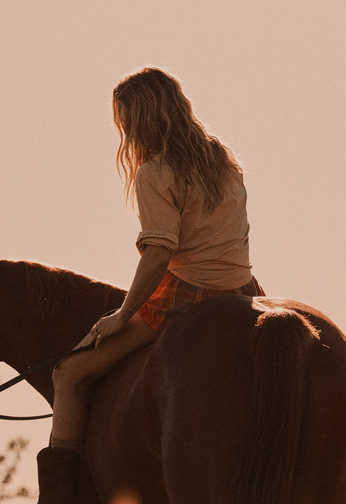 A woman with long light brown hair sits on a horse, shown from behind in warm, backlit outdoor lighting. She wears a beige top and orange plaid shorts against a solid pale peach sky.