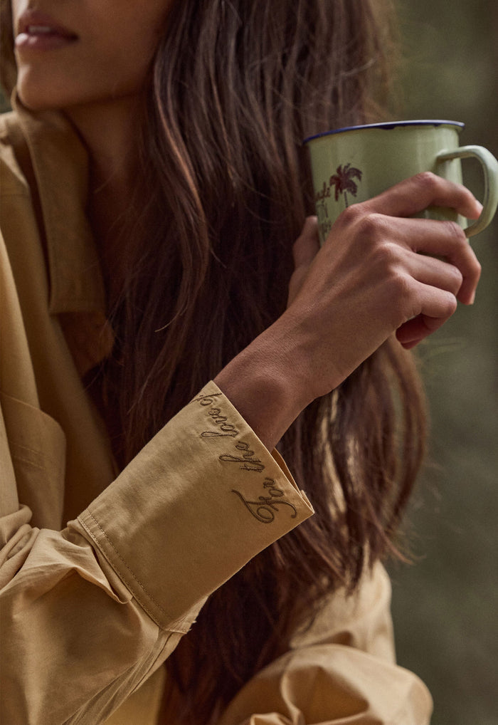 A close-up lifestyle shot of a woman holding a light green enamel mug with a dark rim and palm tree print, wearing a tan button-up shirt with embroidered script on the cuff.