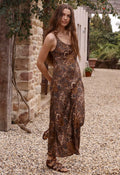 A woman wearing a brown sleeveless maxi dress with a floral pattern, standing on a gravel path next to a rustic stone wall.