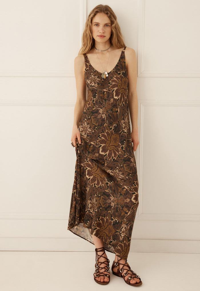 A woman modeling a sleeveless brown maxi dress with a metallic bronze floral pattern, paired with brown strappy sandals, standing against a white paneled wall.