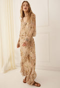 A woman modeling a long-sleeved maxi dress with a beige and brown paisley pattern, standing in a bright, minimalist room with white walls and a cream curtain.