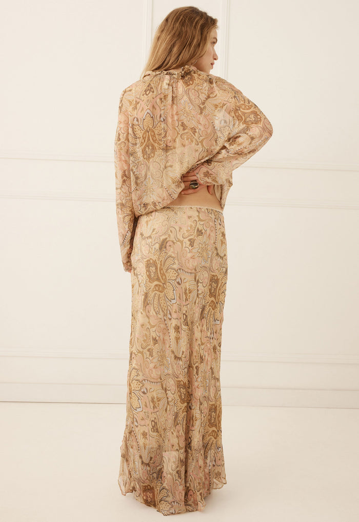A woman stands with her back to the camera wearing a matching two-piece set consisting of a long-sleeve cropped top and a long skirt, both in a beige and brown paisley patterned fabric.