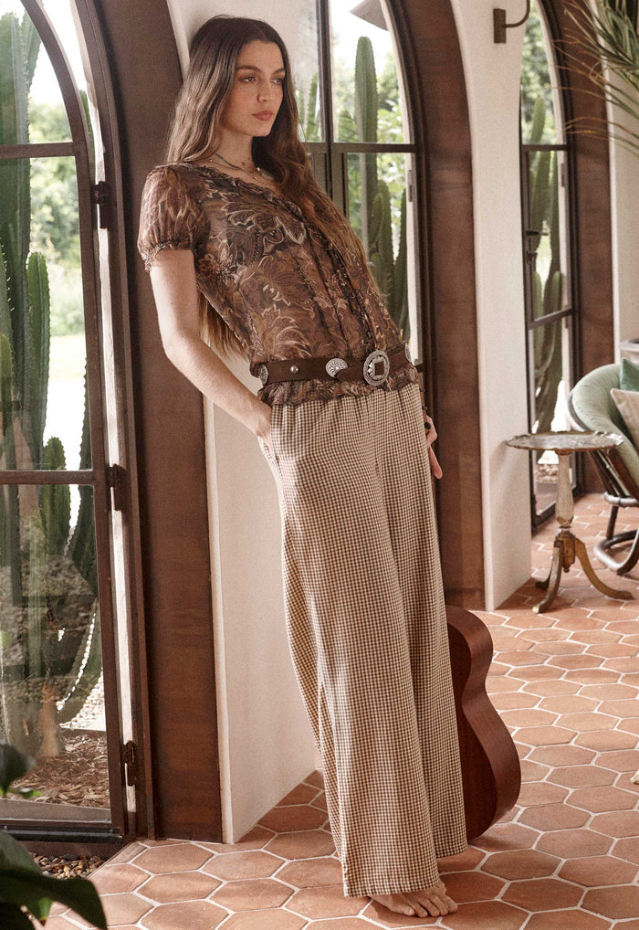 A woman stands barefoot in a rustic indoor setting with arched windows and terracotta tile flooring, wearing a bohemian-style outfit consisting of a brown patterned short-sleeve top, a wide brown belt with large silver conchos, and beige wide-leg checkered pants.