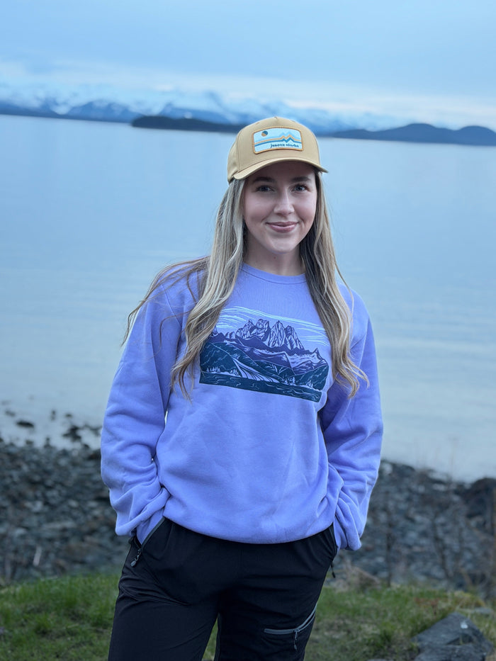 Person wearing a purple sweatshirt with a graphic design, standing by a body of water with mountains in the background.