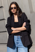 Woman wearing a black blazer and blue jeans standing against a gray wall.
