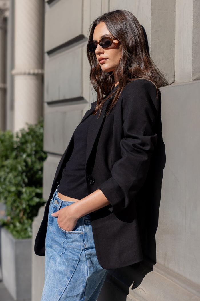 Woman wearing a black blazer and blue jeans leaning against a building.