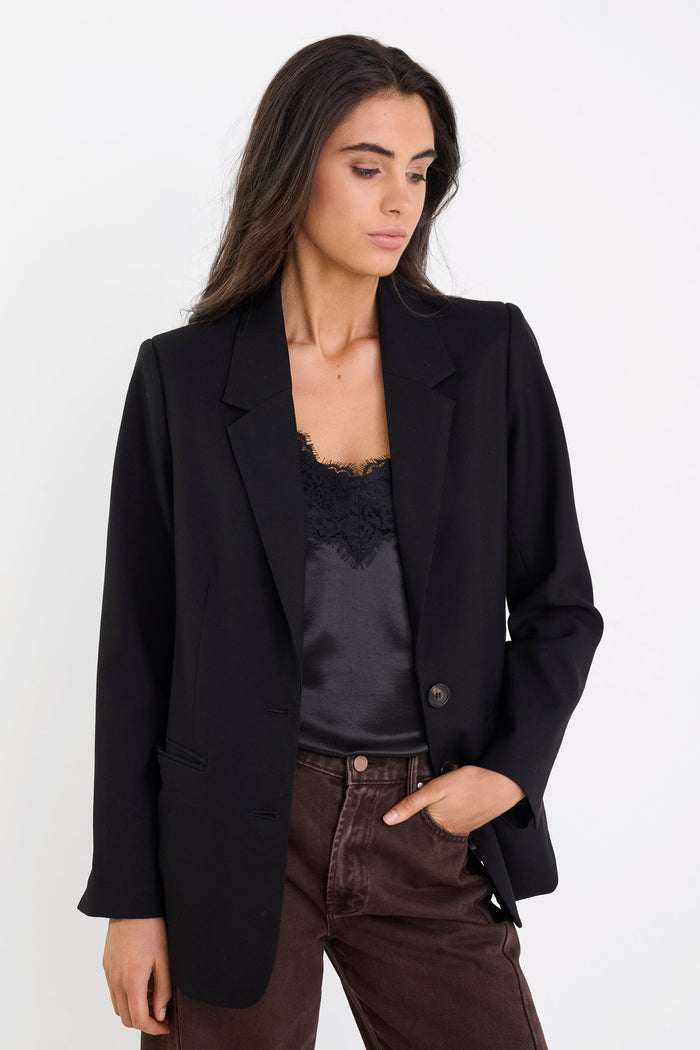 Woman wearing a black blazer over a black top and brown pants on a white background