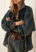 A person wearing an oversized dark blue denim jacket with wide sleeves, paired with a brown patterned top, a brown leather belt with a large silver concho buckle, and blue jeans, set against a white paneled background.