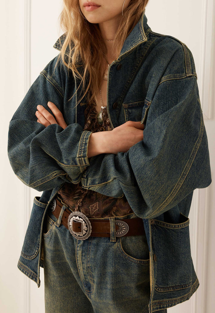 A person wearing an oversized dark blue denim jacket with wide sleeves, paired with a brown patterned top, a brown leather belt with a large silver concho buckle, and blue jeans, set against a white paneled background.