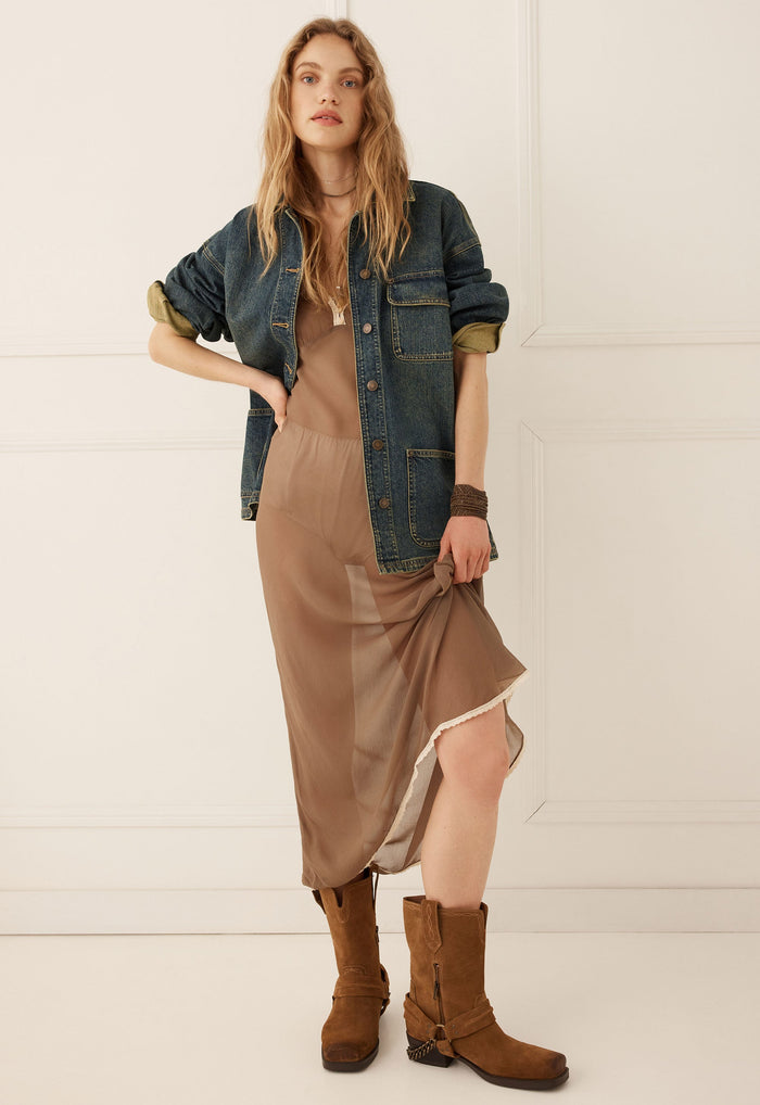A woman models a brown midi slip dress with a sheer overlay, styled with a blue denim shirt jacket, brown leather boots, and a brown choker against a white paneled wall.
