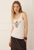 A woman modeling a white tank top with a black winged graphic and a green-and-gold circular emblem on the chest, paired with dark brown pants, against a light cream paneled wall.