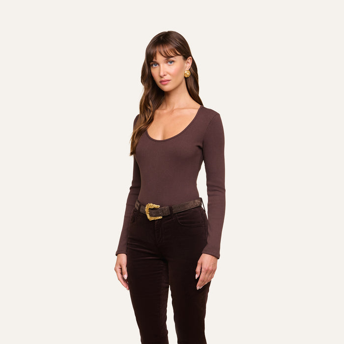 A woman with long brown hair wears the Elara Scoopneck Bodysuit, a fitted, long-sleeve style in brown, paired with dark brown pants, a brown belt with a gold buckle, and gold earrings. She stands against a plain off-white background.