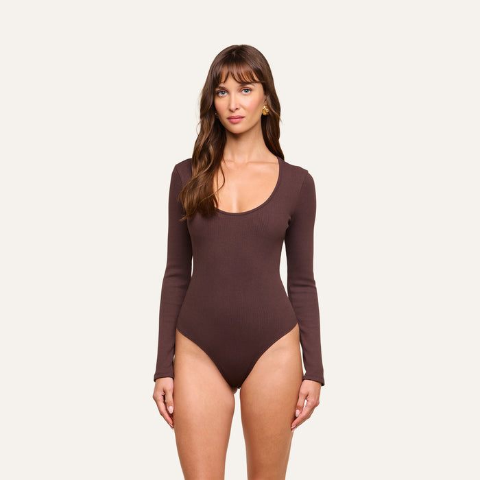 A woman with long brown hair and bangs stands against a plain background, wearing the Elara Scoopneck Bodysuit—a long-sleeve, brown piece crafted from stretch cotton modal for a body-contouring fit. She looks directly at the camera with a neutral expression.