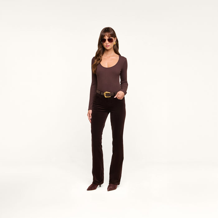 Wearing the Elara Scoopneck Bodysuit in a body-contouring brown hue, paired with matching brown pants and accessorized with brown sunglasses, a woman stands against a plain white background with one hand on her hip.