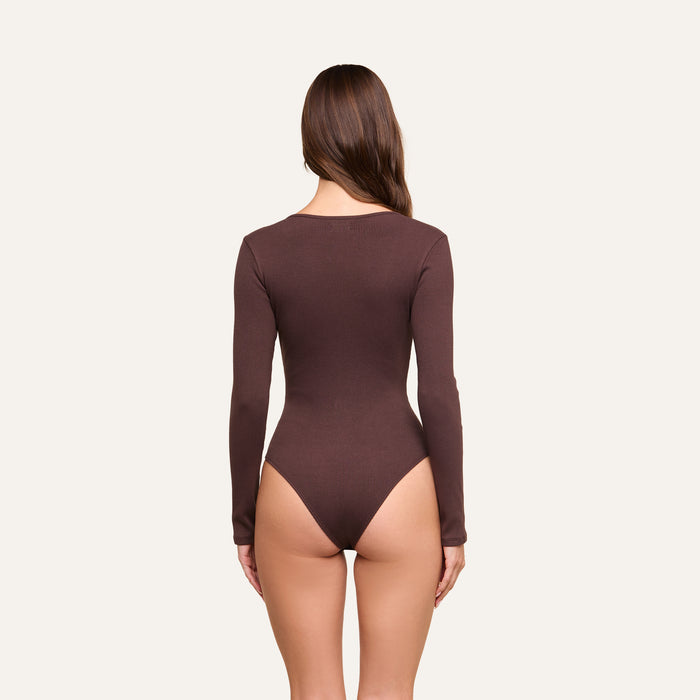 A woman with long brown hair is shown from the back, wearing the Elara Scoopneck Bodysuit—a dark brown, body-contouring, long-sleeve piece crafted from stretch cotton modal—against a plain light background.