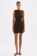 A woman modeling a sleeveless, boat-neck mini dress in a solid dark brown color, paired with dark brown sandals, against a plain white studio background.