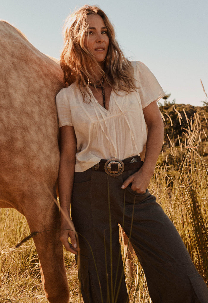 A woman stands outdoors in a field next to a horse, wearing a cream short-sleeve blouse tucked into dark brown wide-leg trousers with a western-style belt and large buckle.