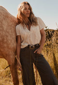 A woman stands outdoors in a field next to a horse, wearing a cream short-sleeve blouse tucked into dark brown wide-leg trousers with a western-style belt and large buckle.