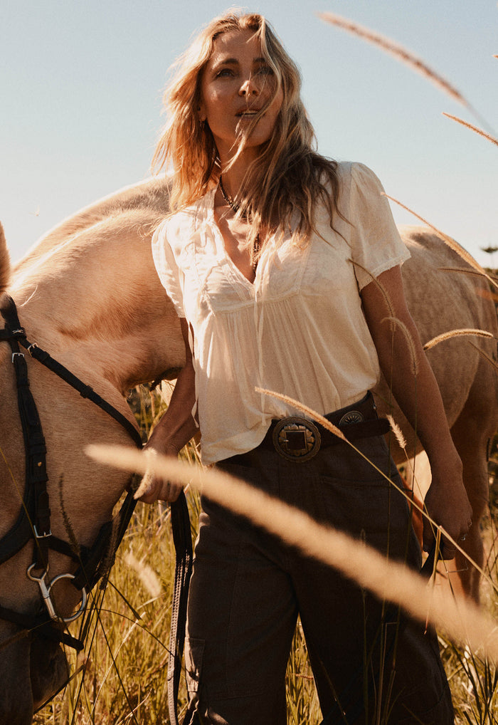 A woman standing outdoors in a field with a horse beside her, wearing a loose white blouse and dark pants with a large round belt buckle.