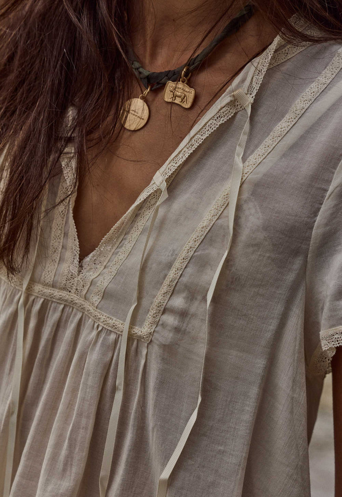 Close-up of a person wearing a loose, short-sleeve bohemian-style top in a light beige or off-white linen-like fabric with lace trim and tie details. The image includes part of the person's face, neck, and hair, and shows a dark cord necklace with wooden pendants.