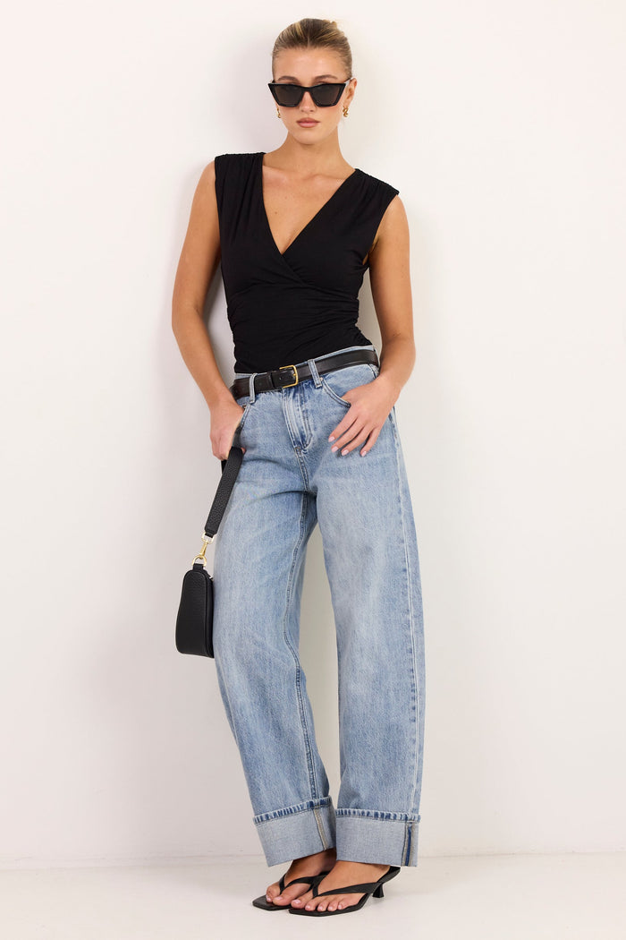 A woman modeling a black sleeveless V-neck top, light-wash wide-leg jeans, black sandals, and sunglasses, holding a small black handbag against a plain white studio background.