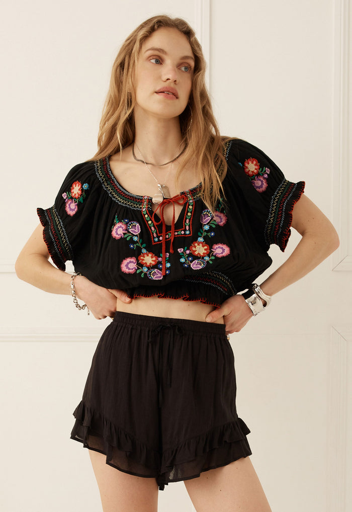 A woman modeling a black short-sleeve crop top with colorful floral embroidery and a red tie detail, paired with black ruffled shorts against a white paneled wall.
