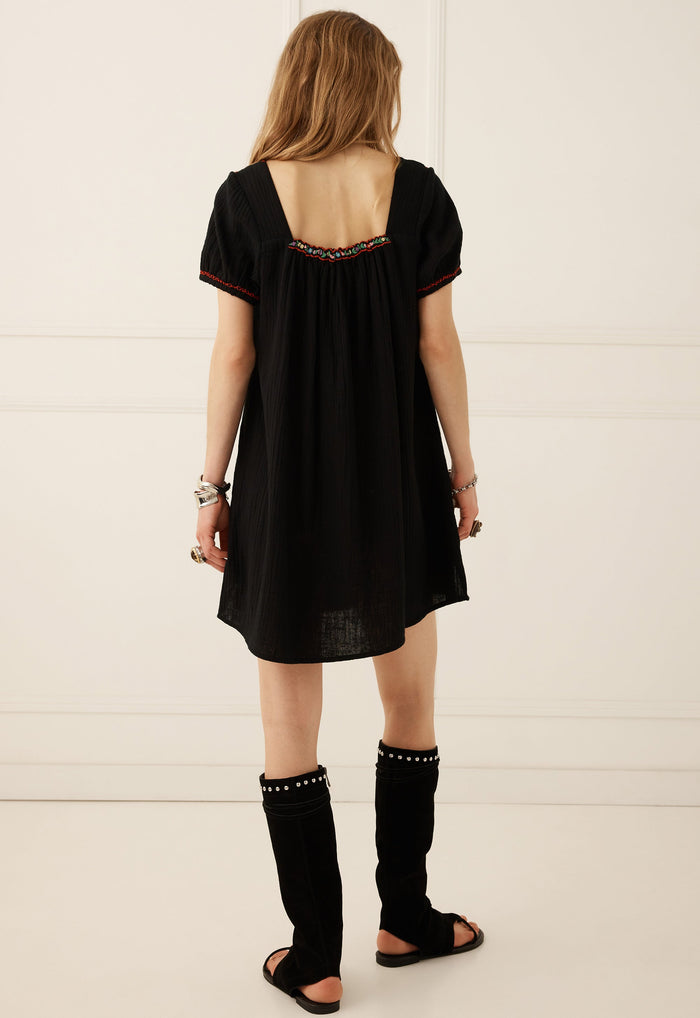 A woman viewed from behind wearing a loose black short-sleeve dress with a decorative embroidered neckline, black studded leg warmers, and black sandals, standing against a white paneled wall.