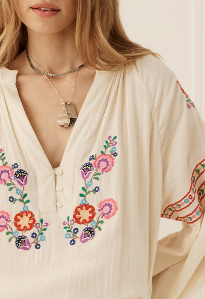 A close-up of a woman wearing a cream-colored bohemian blouse with colorful floral embroidery on the front and sleeves, accessorized with layered silver necklaces.