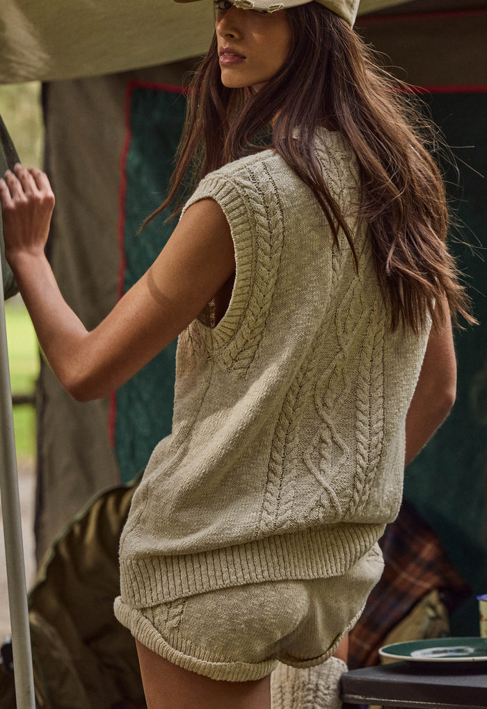 A woman wearing a cream cable-knit sleeveless sweater vest and matching knit shorts, photographed in a rustic outdoor or tent-like setting.