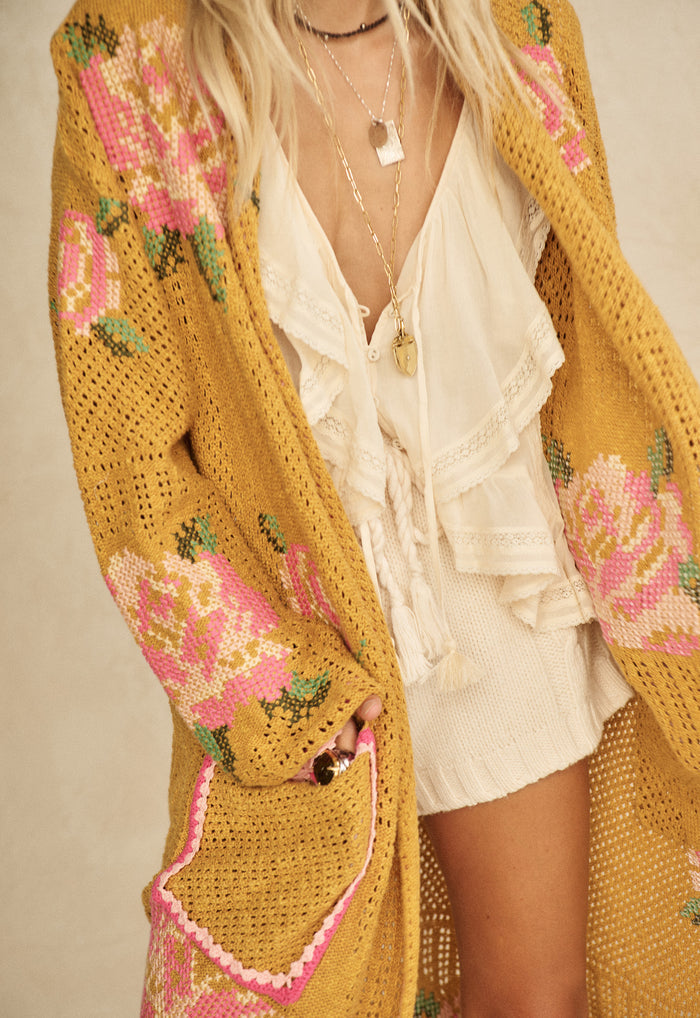 Close-up of mustard floral knit cardigan pocket and open-weave sleeve, styled over cream ruffle blouse
