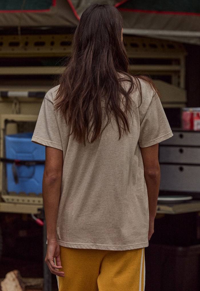 A person viewed from behind wearing an oversized light beige short-sleeve t-shirt and mustard yellow pants, standing in front of a rustic outdoor market stall.